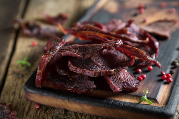 Close-up of sliced beef jerky piled on a wooden serving board with red peppercorns and sprigs of fresh herbs scattered around. The jerky has a rich, dark color and is displayed on a rustic wooden surface.