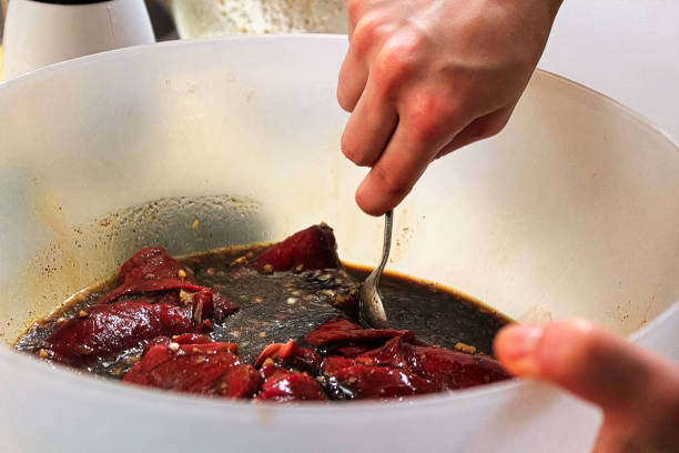 A close-up of raw beef strips being marinated in a bowl of dark liquid seasoning, with a hand stirring the mixture using a spoon. The marinade includes visible spices and herbs, preparing the beef for jerky.